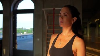 Modern bir spor salonunda büyük bir pencereye doğru ilerleyen azimli bir kızın fotoğrafı. Kendine güveni ve odaklanmış ifadesi fitness ve kişisel hedeflere bağlılığını yansıtıyor..