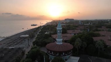 Drone gün batımının sıcak renkleriyle aydınlatılmış bir saat kulesini yakalar. Kule, manzaralı bir plajı, engebeli dağları ve arka planda uzanan sakin denizi olan akşam ışığında dimdik ayakta duruyor.