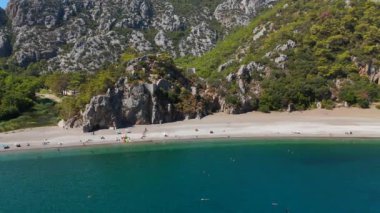 Bu çarpıcı hava manzarası, engebeli kayalık dağlar ve ışıl ışıl Akdeniz tarafından çerçevelenmiş Antalya resimli plajını yakalar. Güneş banyosu yapanlar kıyı boyunca dinlenir, güneşin altında güneşlenirler.