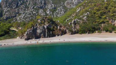 Drone, Antalya 'da görkemli kayalık dağların çerçevelediği güzel bir plajı gösteriyor. İnsanlar yüzerken ve berrak suların tadını çıkarırken görülürler. Doğa ve eğlencenin mükemmel bir karışımını yaratırlar.