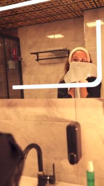 Kadın yıkandıktan sonra yüzünü nazikçe havluyla siler, banyo aynasının önünde durur. Cilt bakımındaki bu ferahlatıcı adım cildinin parlamasını ve kendine bakmasını sağlıyor.,