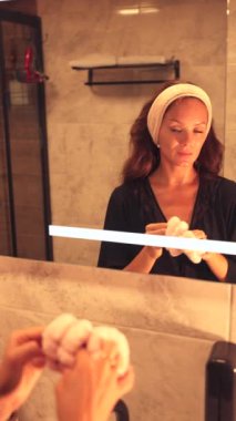 Kadın aynanın önünde duruyor, cilt bakımına hazırlanmak için saç bandı takıyor. Bu küçük ama gerekli adım, saçlarının geri çekilmesini sağlar ve yüz bakımına odaklanmasını sağlar.,