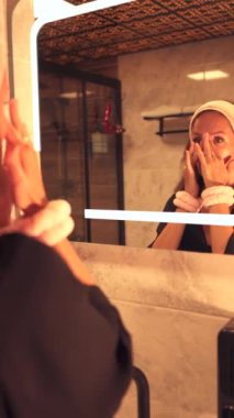 Genç bir kadın banyo aynasının önünde yüzünü nazikçe temizlerken görülüyor. Günlük güzellik rutinini kucaklıyor.. 