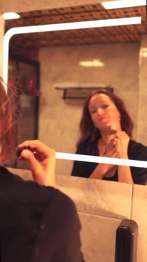 Genç bir kadın banyo aynasının önünde duruyor, kırışıklık gidermek için yüz masajı yapmak için gua sha aleti kullanıyor. Günlük güzellik rutini cildini düzeltmeye ve rahatlatmaya odaklanıyor..