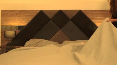 Uzun saçlı esmer bir kadın, rahat ve huzurlu bir yatak odasında yatağını topluyor. Yumuşak ışıklandırma ve sakin atmosfer bir rahatlık ve evde bakım hissini yansıtıyor. Sabah odaklı içerik için mükemmel..
