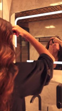 Güzel genç bir kadın banyo aynasının önünde duruyor, alnındaki gua sha aletini günlük güzellik rutininin bir parçası olarak kullanıyor. Rahatlatıcı egzersiz alnın kırışıklık giderilmesine odaklanır.