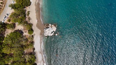 Parlak bir gökyüzü altında doğal arazi ve dağınık yeşillik ile çevrili canlı turkuaz deniz ile kayalık bir sahilin yukarıdan aşağı görüntüsü.. 