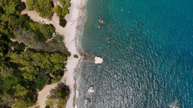 Açık mavi denizle buluşan ormanlık bir kıyı şeridinin hava perspektifi, yemyeşil, engebeli kıyılar ve parlak bir gökyüzünün altında el değmemiş sular sergiliyor.. 