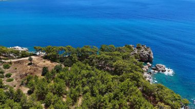 Yoğun çam ormanı ve canlı mavi denizle çevrili engebeli kayalık bir kıyı şeridi parlak bir gökyüzü altında dingin ve doğal bir manzara yaratıyor.. 