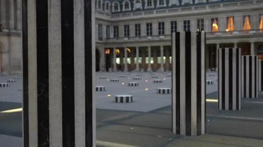Palais Royal 'in aydınlatılmış avlusunda, tarihi mimariyle çevrili çizgili sanat sütunlarının arasında bereli ve ceketli bir kadın yürüyor..