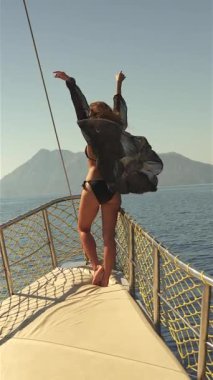 Siyah mayo giyen genç bir kız bir yatın pruvasında zarifçe poz veriyor, arkasında berrak mavi sular uzanıyor.. 