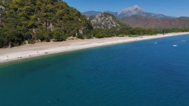Uzaktaki görkemli kayalık dağların çerçevelediği berrak suyla nefes kesici panoramik bir sahil manzarası.