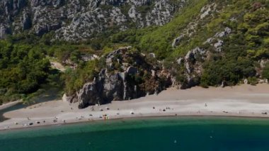 Açık suları ve parlak bir yaz gününde arka planda kayalık dağları olan masmavi bir sahilin panoramik drone görüntüsü.