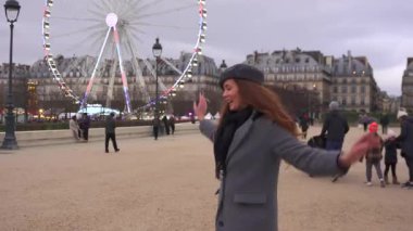 Paris, Fransa 12.20.2024. Gri paltolu ve bereli neşeli bir Fransız kadın şehir meydanında neşeyle dans ediyor. Arka planda dönme dolap var.. 