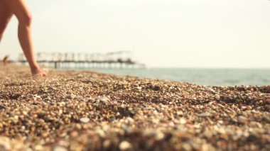 Aydınlık, güneşli bir günde, siyah bir pareo giyerek, suyun yakınındaki boş bir çakıl taşı plajında yürüyen bir kadının bacaklarının yakın çekimi.. 