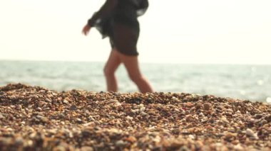 Kıyı şeridi boyunca küçük çakıl taşlarına odaklan. Siyah pareo giymiş genç bir kadın suya doğru yürürken, bulanıklaşır..