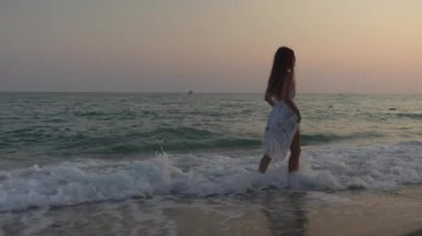 Gün batımında deniz kıyısında gezinen bir kızın sakin sahnesi, sakin okyanus dalgalarından yansıyan altın ışık gibi saçlarını düzeltiyor..