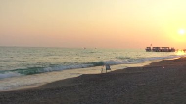 Deniz kıyısında baş döndürücü bir gün batımı. Suyu yansıtan altın gökyüzü, kumlu plaj ve sakin bir atmosfer için yumuşak kamera hareketiyle yakalanmış güneş şezlongları.. 