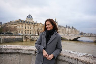 Gülümseyen bir kadın Paris, Fransa 'da bir köprüde duruyor, sıcacık bir paltoyla sarılmış, Seine Nehri' nin tadını çıkarıyor, zarif tarihi binalar, ve şehrin ebedi cazibesi.