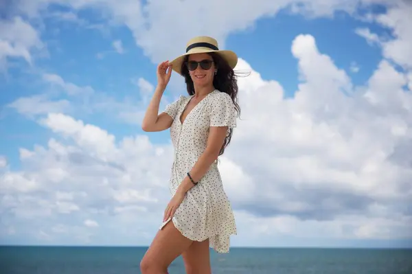 Puantiyeli elbise ve güneş şapkalı neşeli bir kadın okyanusun kenarında duruyor, taze deniz melteminin tadını çıkarırken şapkasını düzeltiyor.. 