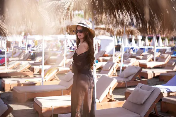 Siyah mayo giymiş şık bir kadın ve saman şemsiyesi altında şezlonglu şezlonglu lüks bir sahil otelinde poz veriyor..