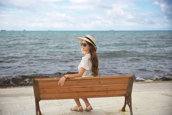 Puantiyeli elbise ve güneş şapkalı genç bir kadın güneşli bir günde, huzurlu okyanus manzarasının tadını çıkarıyor..