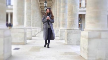 Paris 'te gri paltolu ve bereli bir kız elinde bir fincan kahveyle tarihi sütunlar arasında yürüyor. Kamera hareketi, sahneye dinamik ve modern bir dokunuş ekler..