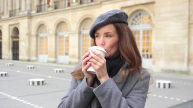 Paris 'te şık bir genç kadının yakın çekimi, gri bereli ve ceketli, rahatlatıcı bir anda kahve fincanı tutan, Fransız yaşam tarzının ve modanın cazibesini yakalayan. 