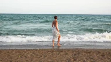 Genç bir kadın gün batımında kumsalda geziniyor, kumların üzerinde yumuşak dalgalar ve okyanusa yansıyan altın ışıkla.. 