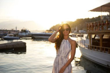 Yazlık elbiseli ve güneş gözlüklü şık bir kadın arka planda tekneleri olan manzaralı bir marinanın yanında altın gün batımının tadını çıkarıyor.. 