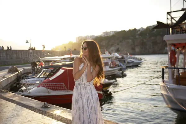 Yaz elbisesi ve güneş gözlüğü takmış neşeli bir kadın, etrafında tekneler ve altın bir gün batımının sıcacık parıltısıyla bir limanın yanında neşe içinde gülümsüyor.. 