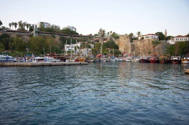 Rıhtım boyunca kenetlenmiş çeşitli tekneleri olan manzaralı bir Akdeniz limanı, tarihi taş duvarlar, yemyeşil yemyeşil ve kıyı kenti arka planı. Antalya. Türkiye 