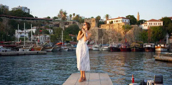 Yazlık elbiseli ve güneş gözlüklü şık bir kadın rıhtımda zarifçe poz veriyor, tekneleri ve tarihi binaları olan manzaralı bir limanda altın günbatımının tadını çıkarıyor..