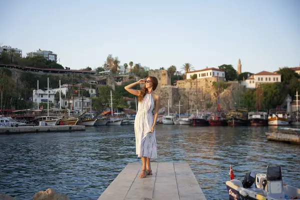 Akan bir yaz elbisesi ve güneş gözlüklü şık bir kadın arka planda tarihi liman manzarası ve tekneleri olan bir rıhtımda güven içinde duruyor. Eski Antalya kenti, Türkiye 