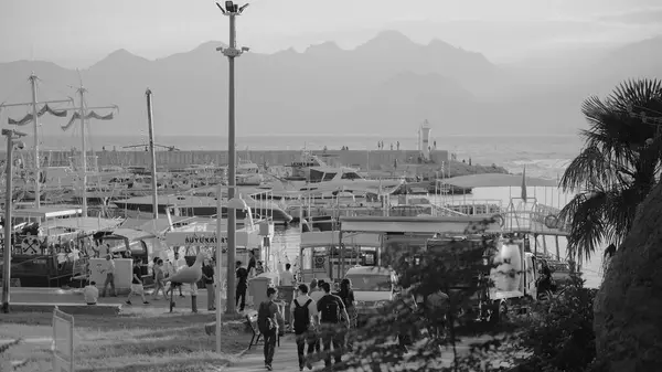Türkiye, Antalya - 29 Eylül 2024: Canlı bir yat limanını teknelerle, yürüyen insanlarla ve deniz kenarında manzaralı bir dağ manzarasıyla gösteren siyah beyaz bir fotoğraf.