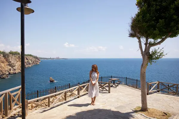 Beyaz bir yaz elbisesi ve güneş gözlüğü takmış bir kadın uçurumun kenarındaki terasta dikilip açık bir gökyüzünün altındaki engin mavi okyanusa bakıyor. Antalya şehrinin Duden Parkı. 