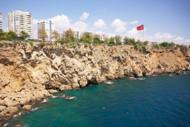 Engebeli kıyı kayalıklarının modern şehir binaları ve Antalya Duden Parkı 'ndaki mavi gökyüzünün altında dalgalanan bir bayrak ile turkuaz denizle buluşması çarpıcı bir manzara. 
