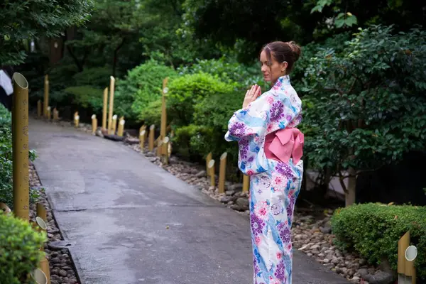 Çiçekli kimono giyen sakin bir kadın Japon bahçesinde elleri dua pozisyonunda, yemyeşil ve dekoratif bambu ışıklarıyla çevrili duruyor..