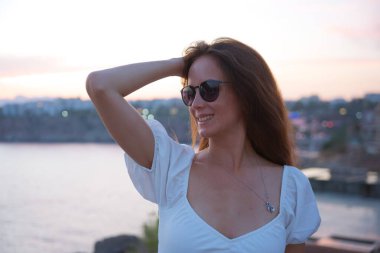 Antalya 'da gün batımında deniz kenarında poz veren uzun saçlı ve güneş gözlüklü mutlu kadın, rahat bir yaz akşamının tadını çıkarıyor.. 