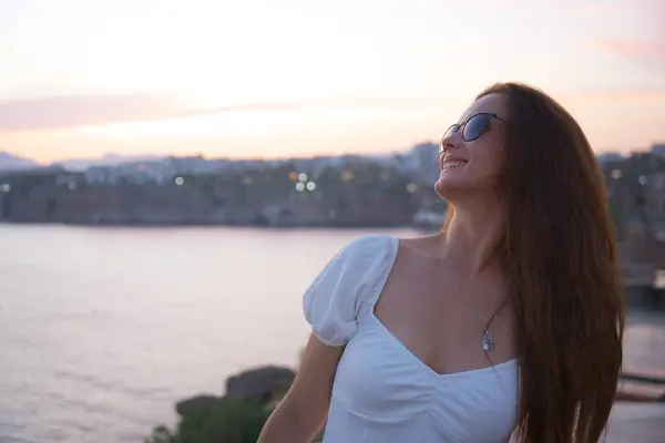 Beyaz bir elbise ve güneş gözlüğü takan gülümseyen kadın, Antalya 'da deniz kenarında huzurlu bir gün batımının tadını çıkarıyor, rahat ve özgür hissediyor.