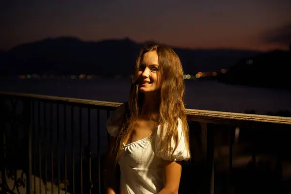 Türkiye, Antalya 'da neşeli bir kadın, deniz kenarında huzurlu bir gecenin tadını çıkarıyor. Güzel bir alacakaranlık, hafif bir aydınlatma ve uzak şehir ışıkları ile sakin bir atmosfer yaratıyor.. 