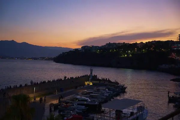 Türkiye 'deki Antalya limanının sakin günbatımı manzarası, kenetlenmiş tekneler, aydınlanmış uçurumlar ve renkli bir akşam gökyüzünün altında dinlenen insanlar