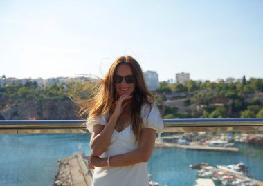 Güneş gözlüklü ve beyaz elbiseli neşeli bir kadın arka planda güzel bir sahil kenti limanı ve marinası olan bir balkonda dikiliyor.. 