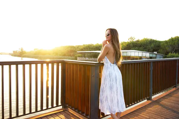 Hafif bir yaz elbisesi içinde gülen kadın ahşap bir terasta duruyor, güneşin batışını seyrediyor doğa ve huzurlu manzarayla çevrili.. 