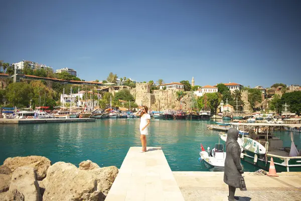 Beyaz elbiseli genç bir kadın bir heykelin yakınındaki iskelede duruyor. Limana bakan tarihi binalar ve tekneler var. Antalya, Türkiye eski kenti