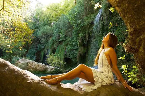 Beyaz elbiseli mutlu bir kadın bir ağaç gövdesinde oturur, yemyeşil, şelale ve altın güneş ışığıyla çevrili, neşeyle arkasını yaslar..