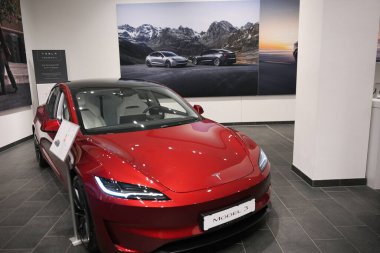 Berlin, Almanya 02.13.2025 Kırmızı Tesla Model 3 elektrikli araç, arka planda diğer Tesla arabalarının yer aldığı tanıtım posterleriyle modern bir galeride sergilendi.. 