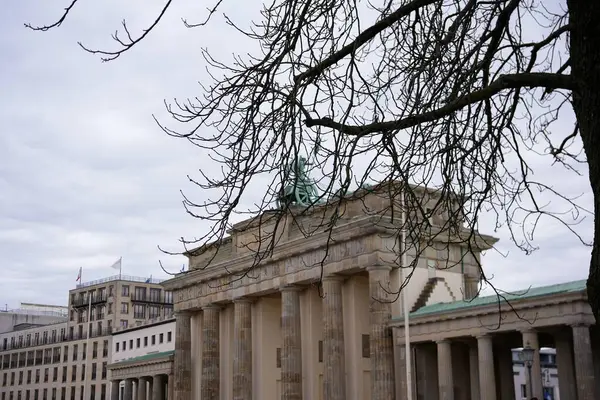 Berlin 'deki Brandenburg Kapısı' nın kısmi bir görüntüsü, arka planda tarihi binalar ve bulutlu bir gökyüzü bulunan çıplak ağaç dalları tarafından çerçevelenmiş..