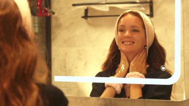 Genç bir kadın günlük cilt bakımı sırasında banyo aynası önünde kozmetik yüz kremi sürerken gülümsüyor.