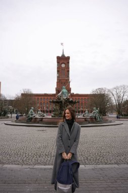 Gri ceketli bir kadın, arka planda kırmızı saat kulesi olan tarihi bir çeşmenin önünde gülümserken sırt çantasını tutuyor.. 